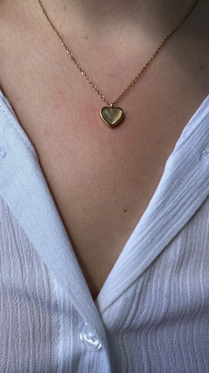 May include: A gold-colored necklace with a heart-shaped pendant. The pendant has a light-colored, possibly mother-of-pearl, center. The necklace is worn against a light blue shirt.