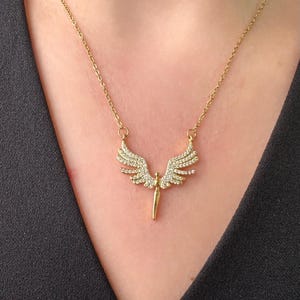 May include: Gold-toned necklace featuring a pendant with an angel wing design. The wings are embellished with small, sparkling stones. The necklace is worn against a black garment, highlighting the jewelry's details.