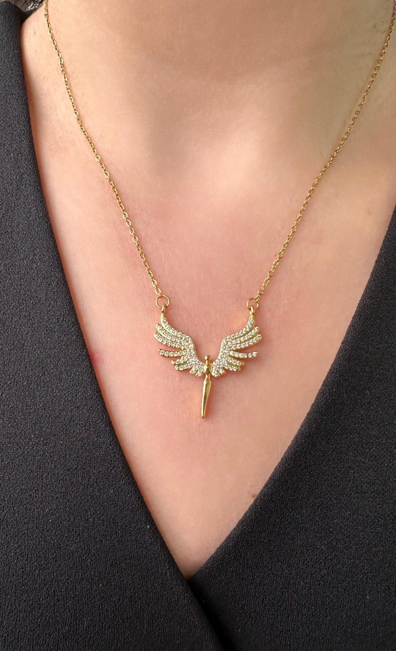 May include: Gold-toned necklace featuring a pendant with an angel wing design. The wings are embellished with small, sparkling stones. The necklace is worn against a black garment, highlighting the jewelry's details.