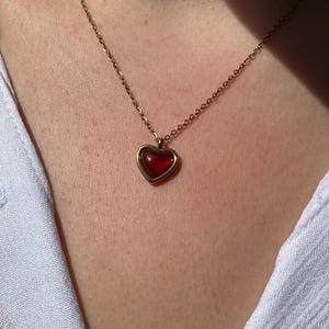 May include: A gold-colored necklace with a heart-shaped pendant. The pendant features a red gemstone set within a gold frame. The necklace is worn against a white shirt.