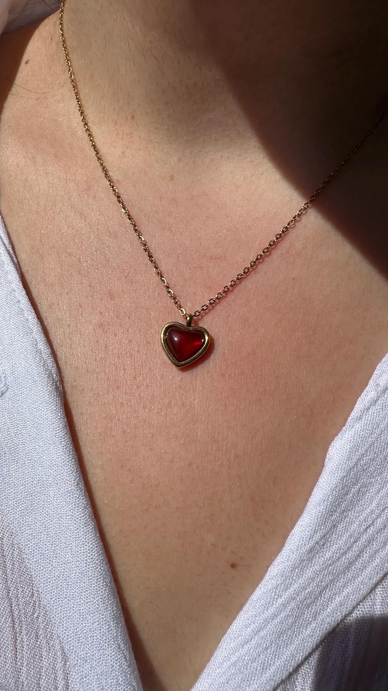 May include: A gold-colored necklace with a heart-shaped pendant. The pendant features a red gemstone set within a gold frame. The necklace is worn against a white shirt.