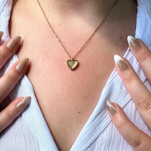May include: A gold-colored necklace with a heart-shaped pendant. The pendant has a light-colored, possibly translucent, center. The necklace is worn over a light blue button-down shirt. The person's hands are visible with long, polished nails.