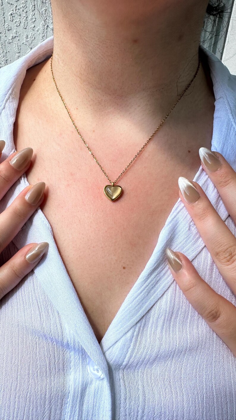 May include: A gold-colored necklace with a heart-shaped pendant. The pendant has a light-colored, possibly translucent, center. The necklace is worn over a light blue button-down shirt. The person's hands are visible with long, polished nails.