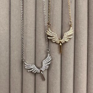 May include: Two necklaces with angel wing pendants. One necklace is silver-toned, and the other is gold-toned. Both pendants feature detailed wing designs with a central figure. The necklaces hang against a neutral, textured background.