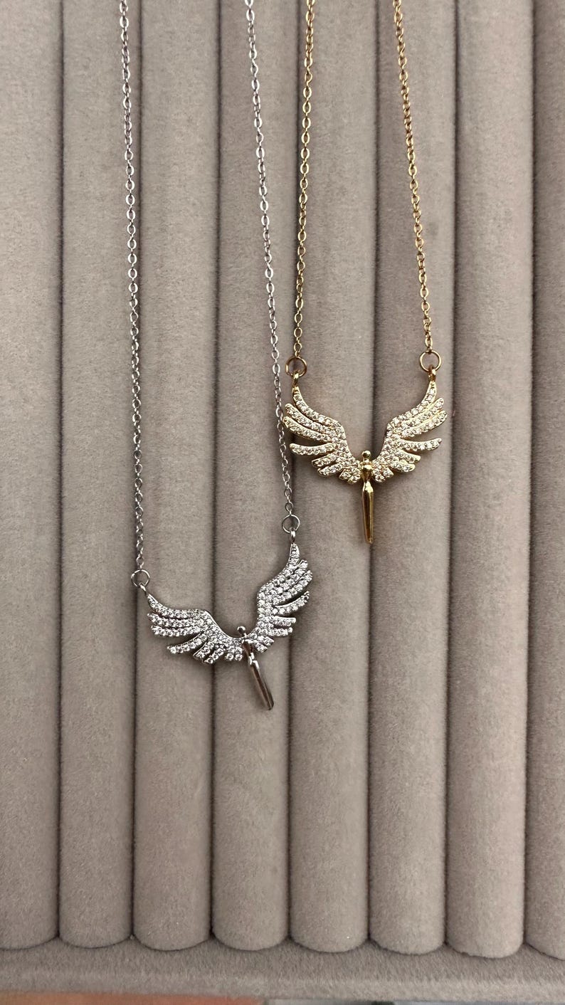 May include: Two necklaces with angel wing pendants. One necklace is silver-toned, and the other is gold-toned. Both pendants feature detailed wing designs with a central figure. The necklaces hang against a neutral, textured background.