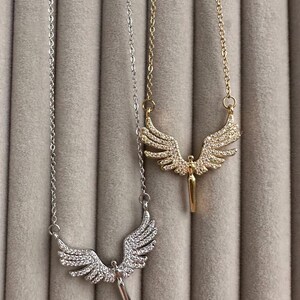 May include: Two necklaces with angel wing pendants. One necklace is silver-toned, and the other is gold-toned. Both pendants feature a detailed wing design with small, sparkling embellishments. The necklaces hang against a soft, gray background.