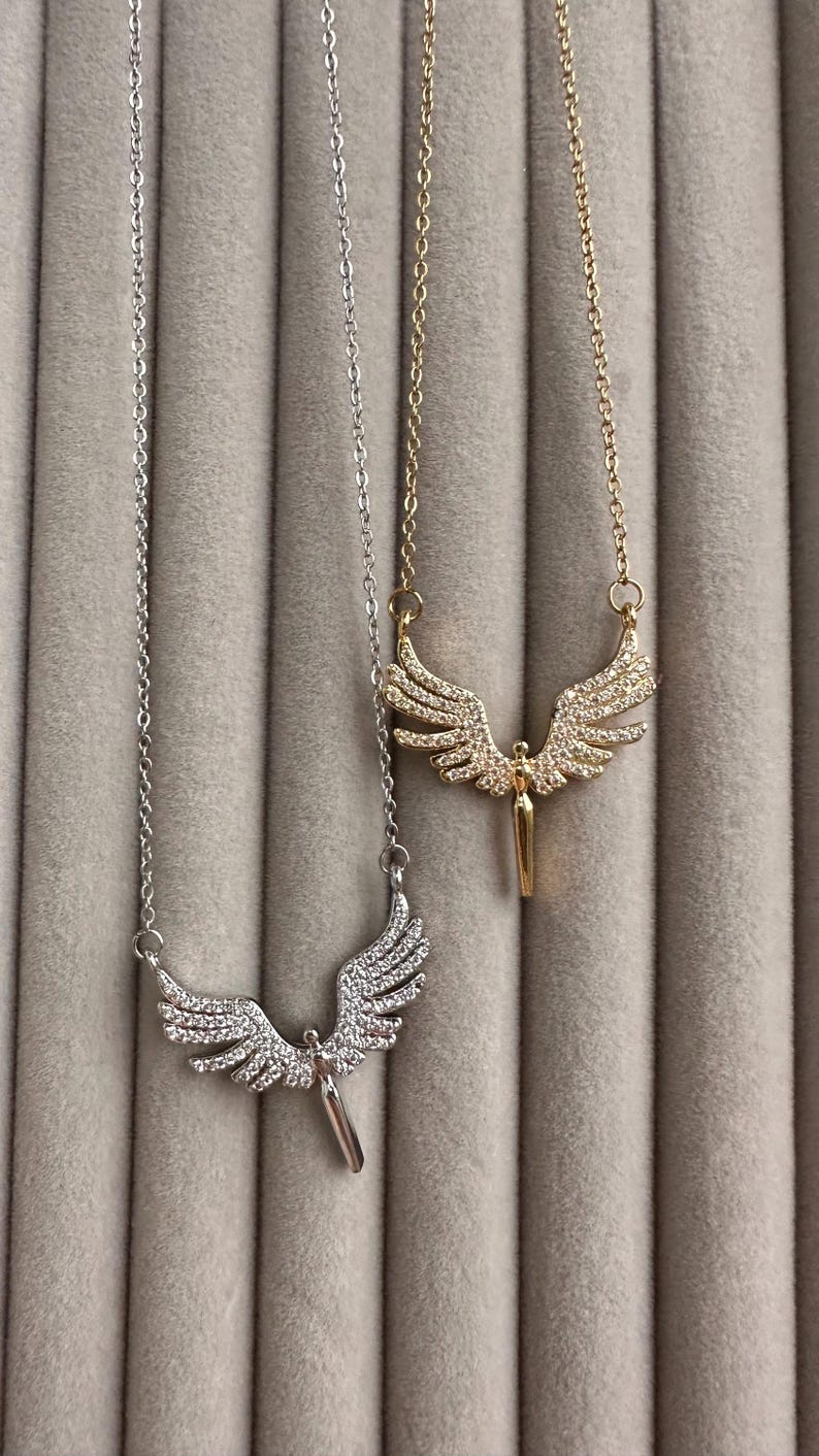 May include: Two necklaces with angel wing pendants. One necklace is silver-toned, and the other is gold-toned. Both pendants feature a detailed wing design with small, sparkling embellishments. The necklaces hang against a soft, gray background.