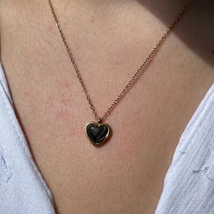 May include: A gold-colored necklace with a heart-shaped pendant. The pendant has a dark, possibly black, center and is framed by a gold border. The necklace is worn against a white shirt.
