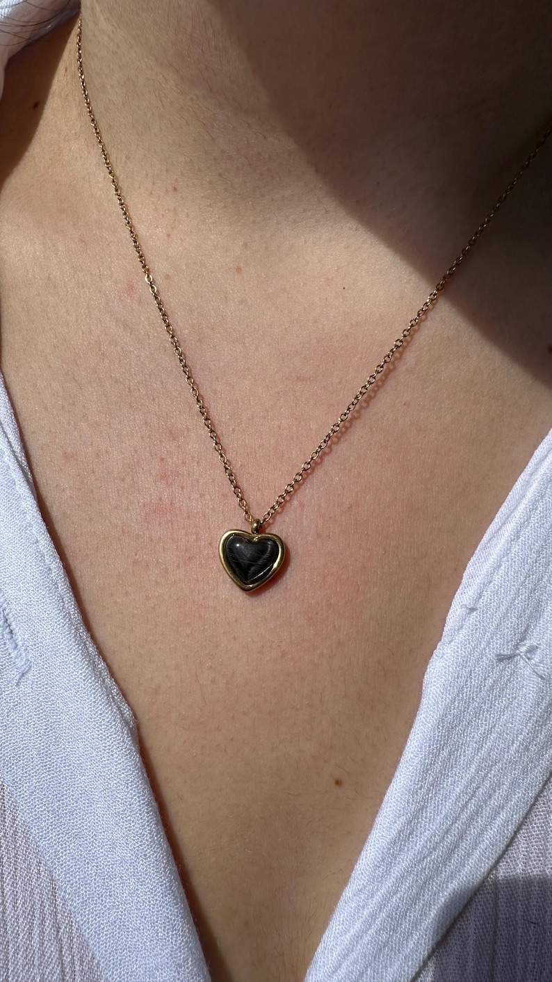 May include: A gold-colored necklace with a heart-shaped pendant. The pendant has a dark, possibly black, center and is framed by a gold border. The necklace is worn against a white shirt.