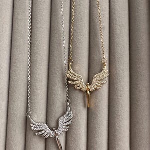 May include: Two necklaces with angel wing pendants. One necklace is silver-toned, the other is gold-toned. Both pendants feature a detailed wing design with small, sparkling embellishments. The necklaces are displayed against a neutral, textured background.