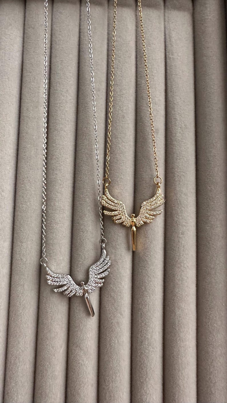 May include: Two necklaces with angel wing pendants. One necklace is silver-toned, the other is gold-toned. Both pendants feature a detailed wing design with small, sparkling embellishments. The necklaces are displayed against a neutral, textured background.