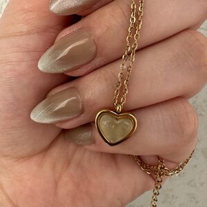 May include: A gold-colored necklace with a heart-shaped pendant. The pendant has a light-colored, possibly mother-of-pearl, center. The necklace is held in a hand with polished nails. The nails are painted with a light brown color.