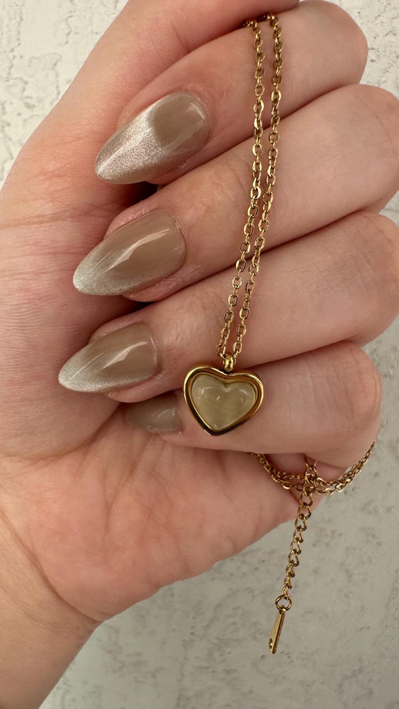 May include: A gold-colored necklace with a heart-shaped pendant. The pendant has a light-colored, possibly mother-of-pearl, center. The necklace is held in a hand with polished nails. The nails are painted with a light brown color.