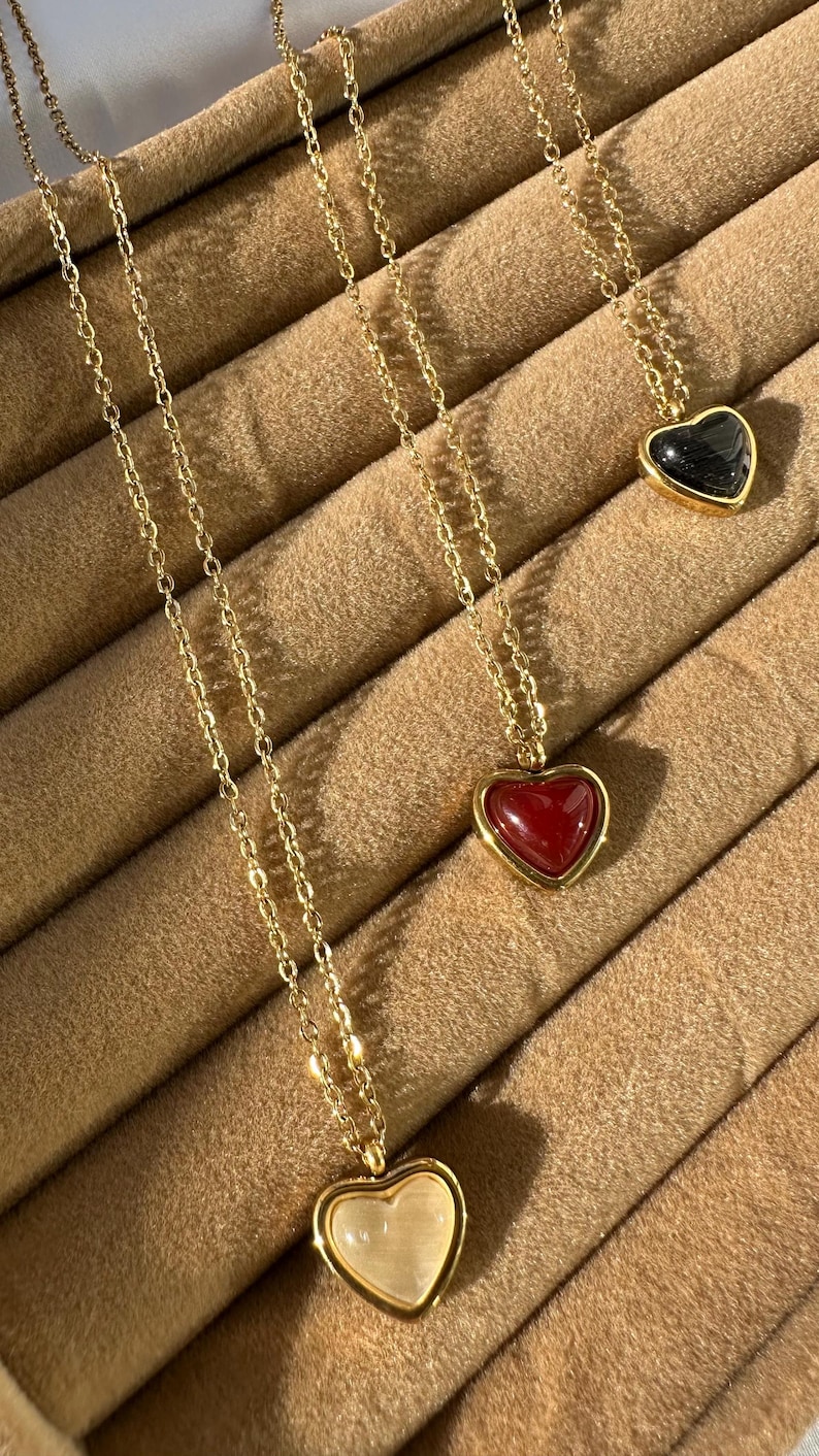 May include: Gold-toned necklaces with heart-shaped pendants in red, black, and cream colors. The pendants are set in gold frames and hang from delicate chains. The necklaces are displayed on a brown surface.