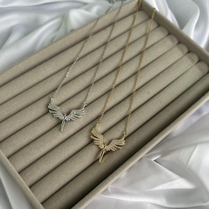 May include: Two necklaces with angel wing pendants. One necklace is silver-toned, the other is gold-toned. Both pendants feature a detailed wing design with small, sparkling embellishments. The necklaces are displayed on a beige surface.