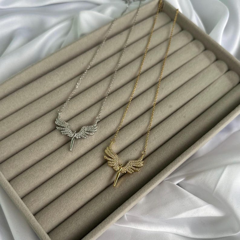 May include: Two necklaces with angel wing pendants. One necklace is silver-toned, the other is gold-toned. Both pendants feature a detailed wing design with small, sparkling embellishments. The necklaces are displayed on a beige surface.