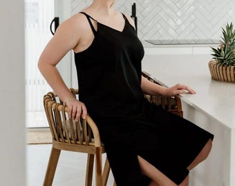 May include: A black slip dress with spaghetti straps and a side slit. The dress is knee-length and worn by a person sitting on a wooden chair. The chair has a natural wood finish. The background includes a kitchen with white cabinets and a pineapple.