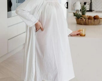 May include: A white cotton robe and nightgown set. The long-sleeved robe has a relaxed fit, and the nightgown features a square neckline. The scene is set in a bright, modern kitchen with a woven pendant light and a white countertop.
