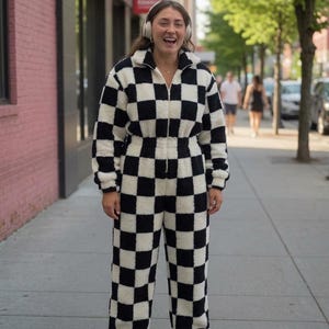 May include: A black and white checkered jumpsuit with a zip-up front. The jumpsuit is made of a soft, fuzzy material. The person is wearing white headphones and sneakers. The background includes a brick building and trees.