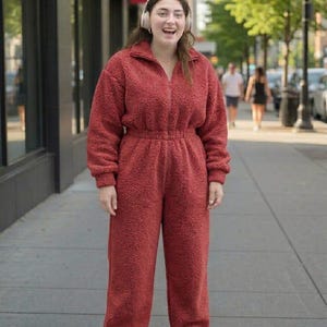 May include: A rust-colored, long-sleeved, one-piece jumpsuit made of a textured, fuzzy fleece material. The garment features a high collar, a full-length front zipper, an elasticized waistline, and long, straight-leg trousers with ribbed cuffs.