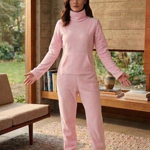 May include: A light pink fleece jumpsuit featuring a high turtleneck collar, long sleeves, and a front kangaroo pocket. The one-piece garment has full-length pants that extend into integrated foot coverings, providing a uniform look in soft fabric.