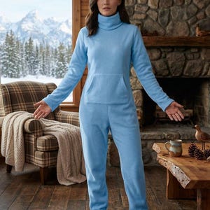 May include: A light blue fleece jumpsuit featuring a high turtleneck collar, long sleeves, and a front kangaroo pocket. The one-piece garment includes integrated foot coverings, providing full-body coverage in a soft, solid-colored fabric.