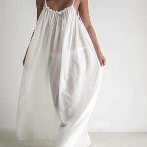 May include: A flowing, white maxi dress with thin spaghetti straps and a gathered neckline. The lightweight fabric drapes loosely, creating a relaxed silhouette. The dress is floor-length and suitable for warm weather.