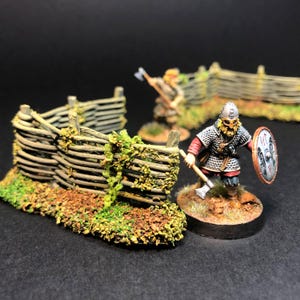 Peut inclure: Figurine miniature de guerrier viking avec une hache et un bouclier, près d'une clôture en bois. La figurine porte une armure en cotte de mailles et un casque. La clôture est faite de planches de bois et couverte de feuillage vert.