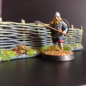 Peut inclure: Figurine miniature d'un guerrier viking peint à la main, avec une armure en cotte de mailles détaillée, un casque et une lance. La figurine est posée devant une clôture en osier et un terrain herbeux. La palette de couleurs comprend des nuances de marron, bleu, rouge et argent métallique.