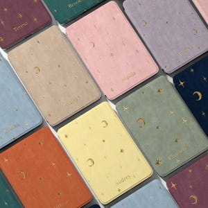 Puede incluir: Una colección de fundas rectangulares en varios colores, incluyendo burdeos, verde azulado, beige, rosa, lavanda, azul claro, amarillo, naranja y azul oscuro. Cada funda presenta un patrón de lámina dorada de estrellas y lunas.