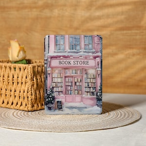 Pode incluir: Capa retangular rosa e branca com uma ilustração de uma livraria no inverno. A imagem mostra uma cena de inverno com neve e árvores. A placa acima da loja diz "BOOK STORE".