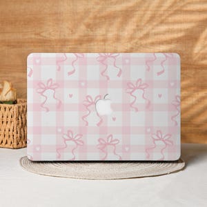 Könnte beinhalten: Eine Laptop-Hülle mit rosa-weißem Vichy-Muster, verziert mit rosa Schleifen und kleinen Herzen. Das Apple-Logo ist sichtbar. Die Hülle liegt auf einem runden, gewebten Platzset.