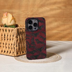 May include: A smartphone case with a dark red background and a pattern of black bats. The case has a black border and a camera cutout. The case is sitting on a round, woven placemat.