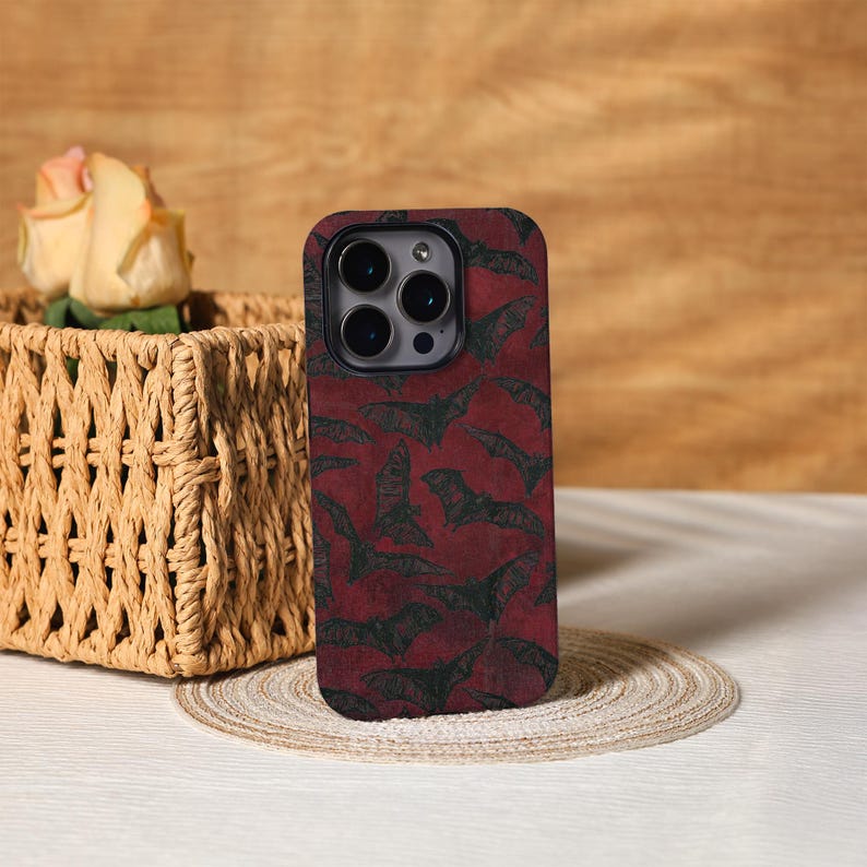 May include: A smartphone case with a dark red background and a pattern of black bats. The case has a black border and a camera cutout. The case is sitting on a round, woven placemat.