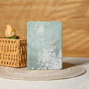 以下が含まれることがあります： 長方形の淡い緑色の電子書籍リーダーカバー。白い桜の花が右下に集まり、上部に散らばっています。カバーは、バスケットとバラを背景にした織りのプレースマットの上にあります。