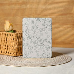 May include: An off-white rectangular case with a pattern of birds and floral branches in muted green. The case is placed on a woven placemat, with a wicker basket and a yellow rose in the background.