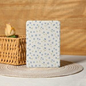 Puede incluir: Una funda rectangular para tableta adornada con un delicado estampado floral de pequeñas flores azules y tallos verdes. La funda está sobre un mantel individual tejido, con una cesta de mimbre y una rosa amarilla en el fondo.