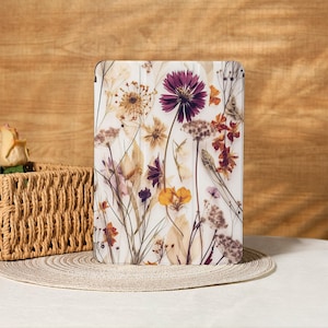 Puede incluir: Una funda blanca para tableta con un diseño de flores prensadas. El arreglo floral incluye flores moradas, amarillas y naranjas con delicados tallos y hojas. La funda está sobre un fondo de madera clara.