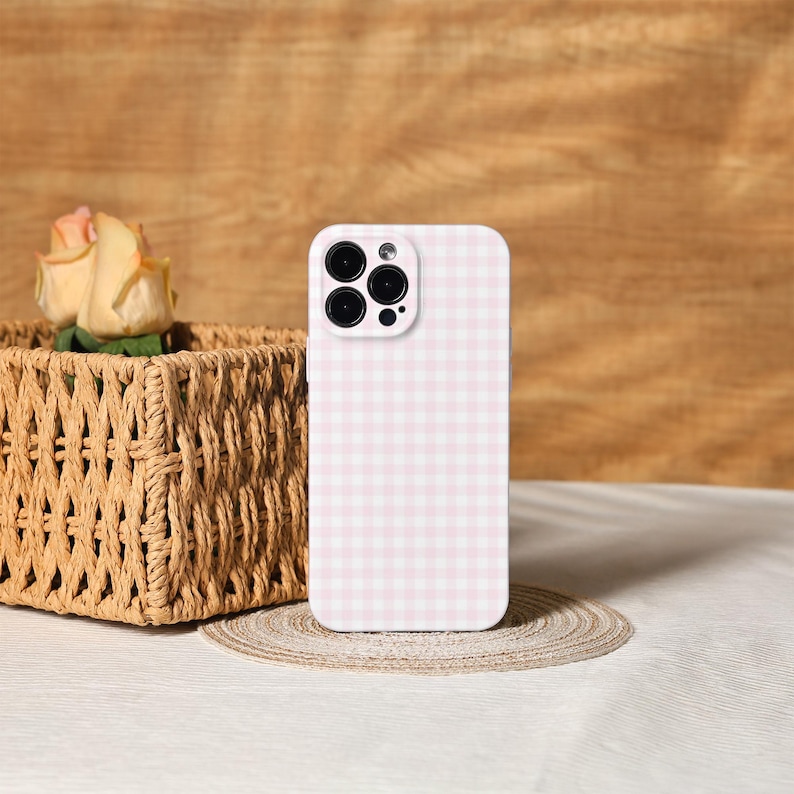 May include: A pink and white gingham patterned phone case. The case is standing upright on a small, round, woven mat. A woven basket with a yellow rose is in the background.