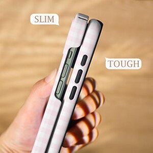 May include: Two phone cases, one partially inserted into the other, are shown in a side view. The cases are light pink with a white gingham pattern. The word "SLIM" is in a white speech bubble. The word "TOUGH" is in a white speech bubble.