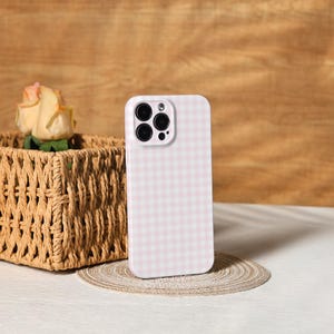 May include: A pink and white gingham patterned phone case. The case is standing upright on a small, round, woven mat. A woven basket with a rose is in the background.