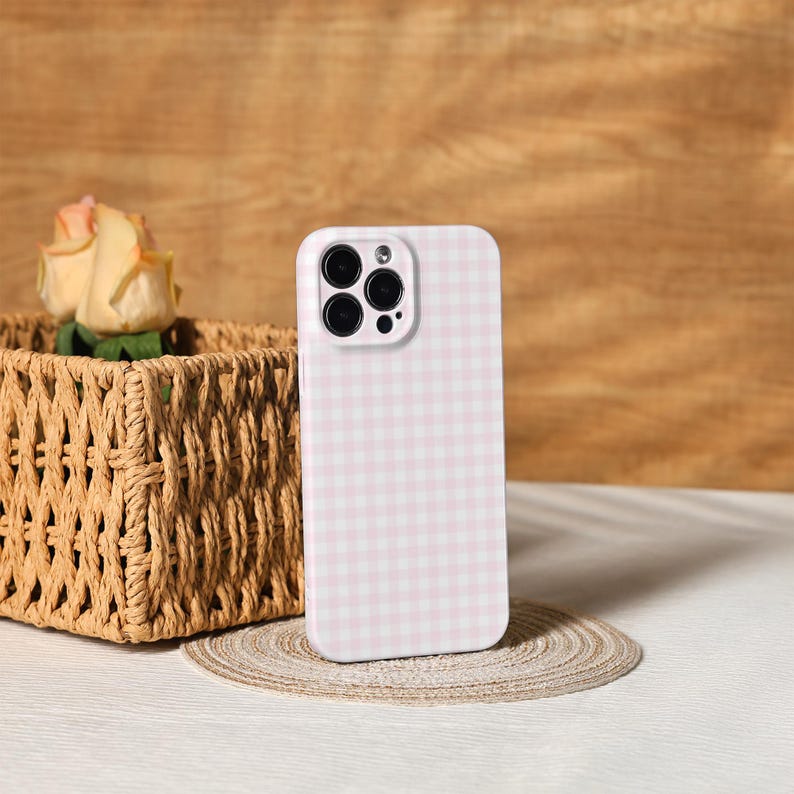 May include: A pink and white gingham patterned phone case. The case is standing upright on a small, round, woven mat. A woven basket with a rose is in the background.