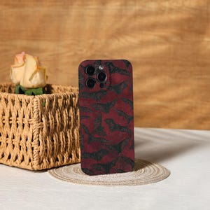May include: A smartphone case with a dark red background and a repeating pattern of black bat silhouettes. The case has a glossy finish and features a camera cutout. The case is displayed on a neutral surface.