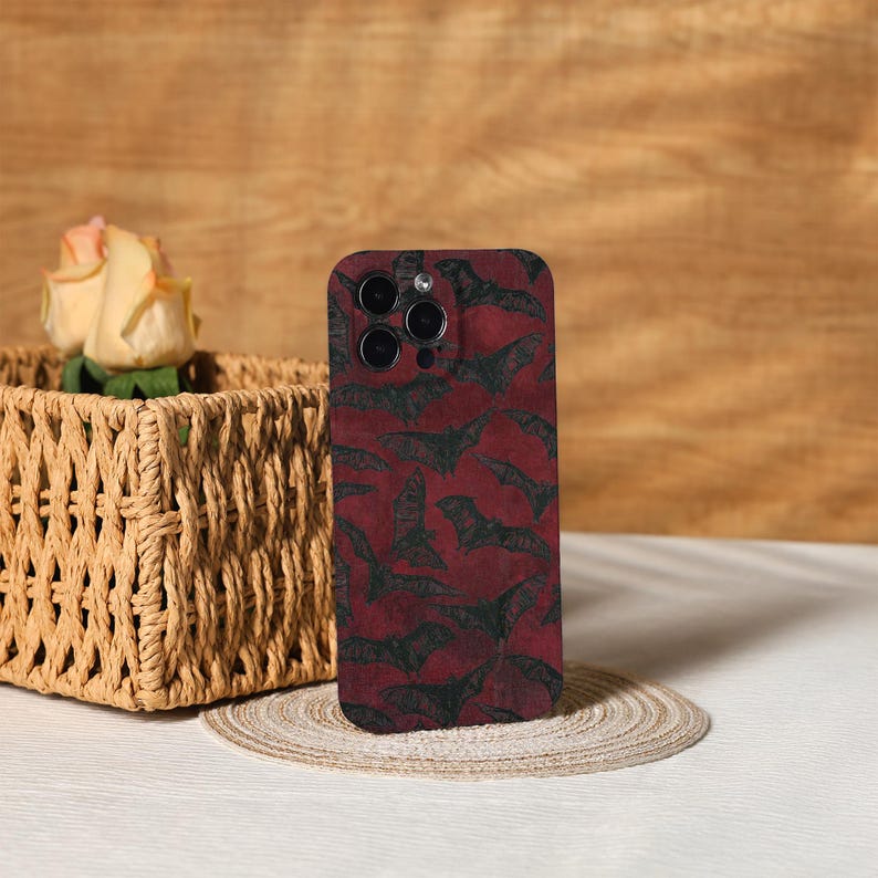 May include: A smartphone case with a dark red background and a repeating pattern of black bat silhouettes. The case has a glossy finish and features a camera cutout. The case is displayed on a neutral surface.