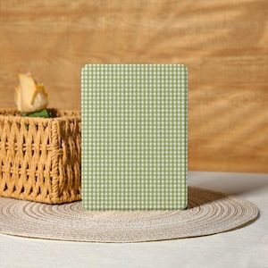 Puede incluir: Un objeto rectangular con esquinas redondeadas, con un patrón de cuadros verdes y blancos. Una cesta tejida con una rosa y un salvamanteles tejido están en primer plano. El fondo es una superficie de madera marrón claro.