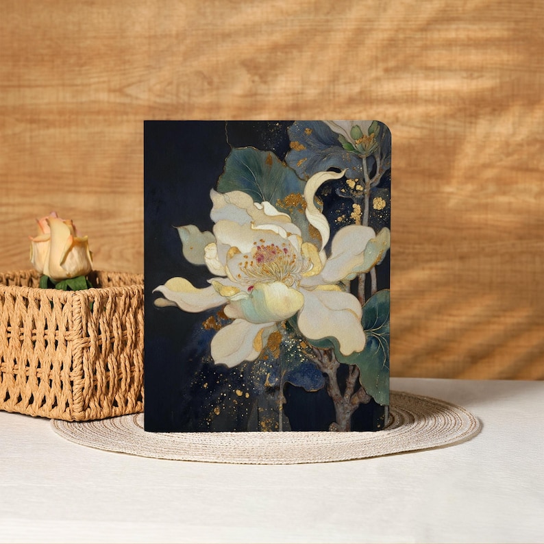 May include: A floral notebook featuring a large white flower with yellow and green accents against a dark blue background. The design includes gold flecks and green leaves. The notebook is displayed on a neutral surface.