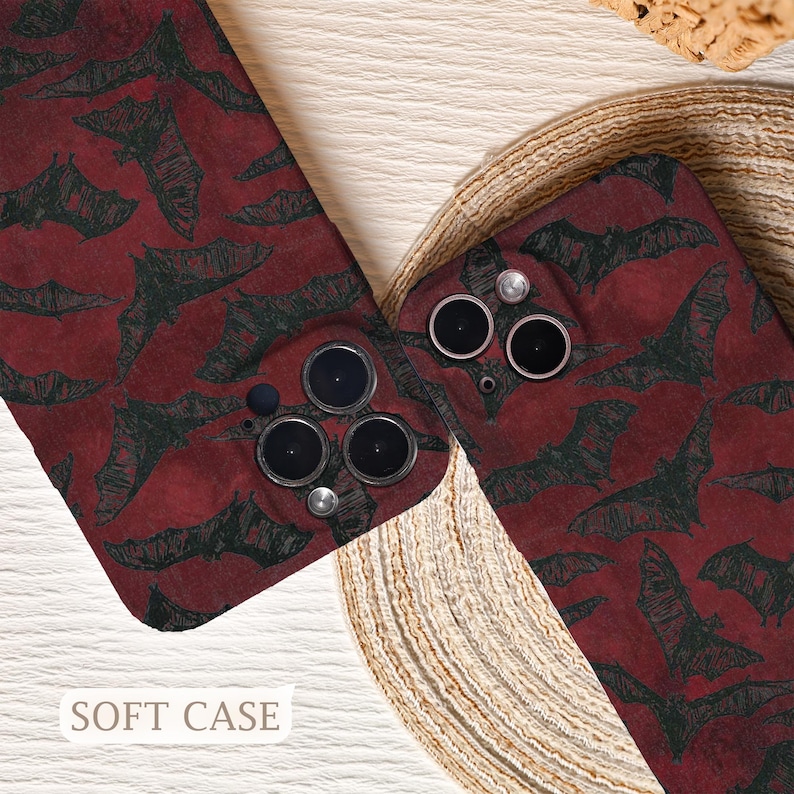 May include: Two phone cases with a repeating pattern of black bats on a deep red background. The cases have camera cutouts and the text "SOFT CASE" is visible at the bottom. The cases are displayed on a woven surface.