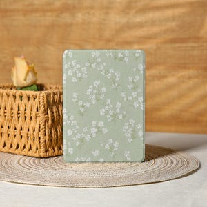 May include: A light green e-reader cover with a delicate white floral pattern. The cover is rectangular and features a repeating design of small white flowers and green leaves. The background is a soft, muted green. The cover is on a neutral surface.