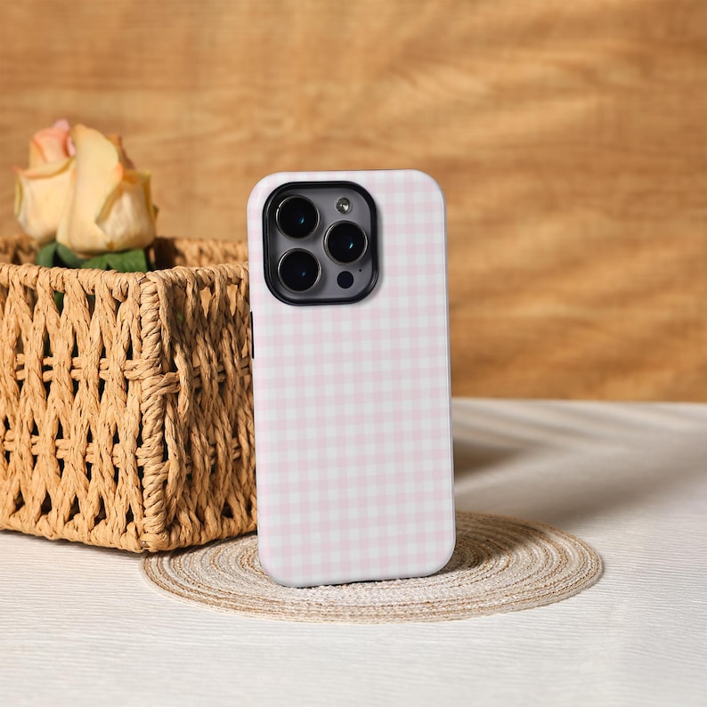 May include: A pink and white gingham patterned phone case. The case is standing upright on a small, round, woven placemat. The phone case has a black camera lens and a black border. A woven basket and a rose are in the background.
