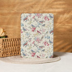 May include: A beige tablet case featuring a floral and bird pattern. The design includes pink, blue, and red flowers with various birds perched on branches. The case is rectangular and has a smooth surface.